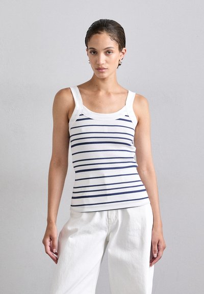 Weißes ärmelloses Tanktop mit navyblauen, horizontalen Streifen, gerippter Textur und Rundhals-Ausschnitt. Kombiniert mit lockeren weißen Hosen.