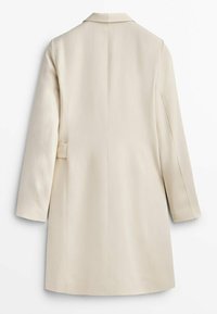 Manteau long beige avec manches droites, un col et une couture dans le dos, montré de dos sur fond blanc.