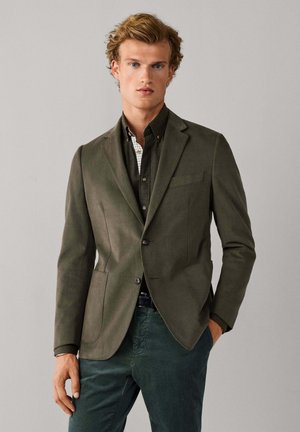 Jonge man met een olijfgroene blazer, donkergroen overhemd en groene broek, staand met één hand in de zak tegen een effen achtergrond.