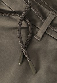 Tissu vert olive avec une surface texturée et un cordon tressé noir, présentant des embouts en caoutchouc pour les ajustements et une taille soigneusement cousue.