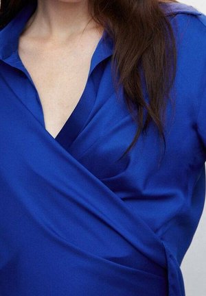 Blouse bleue à col en V profond, fabriquée en tissu lisse, avec des manches longues et un drapé doux sur la poitrine.