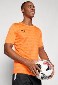 Orange kurzärmliges Sportshirt mit strukturiertem Muster, schwarzem Puma-Logo und rundem Ausschnitt, kombiniert mit einem Fußball in der Hand.