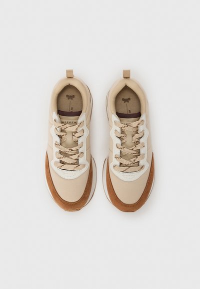 Beige and brown sneakers featuring a canvas upper, suede accents, rounded toe, and textured laces, with a pull tab at the heel.