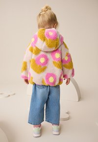 Veste à capuche en polaire avec motifs floraux roses et jaunes, associée à un jean denim à jambes larges et des baskets pastel. Texture douce et couleurs vives.