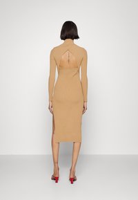 Abito beige a coste, con maniche lunghe e collo alto, schiena scoperta e spacco laterale, abbinato a sandali rossi con tacco. Texture liscia.