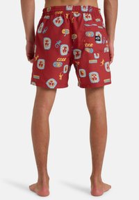 Piros fürdőshorts, állítható derekú, színes arcokkal, gyíkokkal és pálmafákkal díszített mintázattal, könnyű anyagból készült.