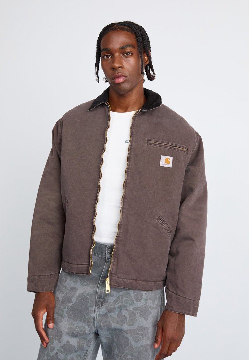 Bruine zip-up jas met een zwarte kraag, voorzien van een klein logo op de borst, gecombineerd met een grijs met patroon broek en een wit shirt eronder.