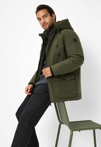 Giacca isolante verde scuro con cappuccio, dotata di tasche con zip e una tasca frontale con patta. Indossata sopra una maglietta grigia chiara e pantaloni neri.