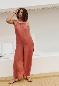 Jumpsuit boje ružićaste patine od lana s halter vratom i širokim nogavicama, s naboranim dekolteom, u kombinaciji s ravnim sandalama. Teksturirana pozadina.