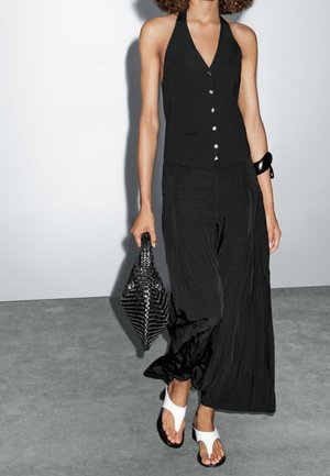 Woman wearing sleeveless black buttoned top and loose black pants, carrying a textured black clutch, wearing white sandals and black bracelet.