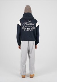 Námornícka mikina s bielymi akcentmi a vyšívaným textom na zadnej strane; v kombinácii s svetlosivými nohavicami a béžovými topánkami. Wľnová pletená čiapka.