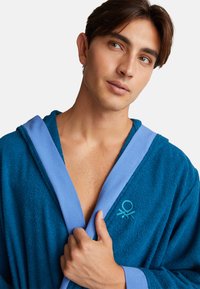 Jeune homme aux cheveux bruns portant un peignoir bleu, tenant le col d'une main, regardant légèrement sur le côté.