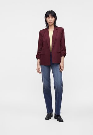Kvinde stående mod en ensfarvet baggrund iført en bordeauxfarvet blazer, gul top, blå jeans og sorte ankelstøvler med hæl.