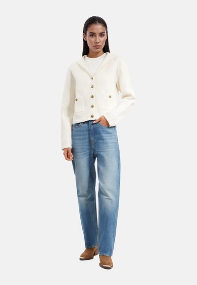 Hvid hætte cardigan med gyldne knapper, to forlommer; parret med lyseblå lige ben jeans og brune hælede støvler.