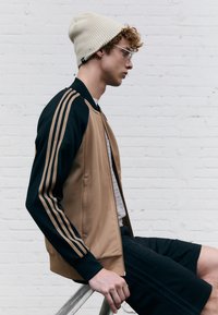 Ruskea ja musta urheilutakki, jossa on kolme kultaraitaa hihoissa, päällä harmaa paita ja mustat shortsit, yhdistetty beigeen pipoon.