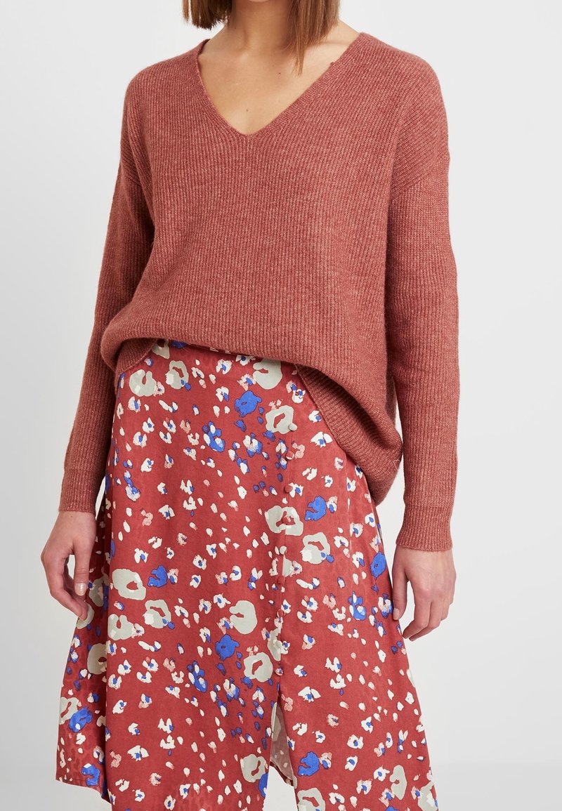Femme portant un pull en maille couleur rouille à manches longues avec un col en V, rentré dans une jupe midi fleurie rouge aux motifs bleus, beiges et blancs.