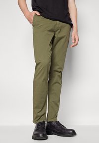 Pantaloni di cotone verde oliva con vestibilità slim, dotati di chiusura con bottone e zip, tasche laterali, indossati con scarpe nere stringate.