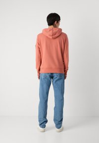 Felpa con cappuccio in cotone rosa con polsini e orlo a coste, abbinata a jeans di denim blu chiaro e sneakers bianche. Vista dal retro.