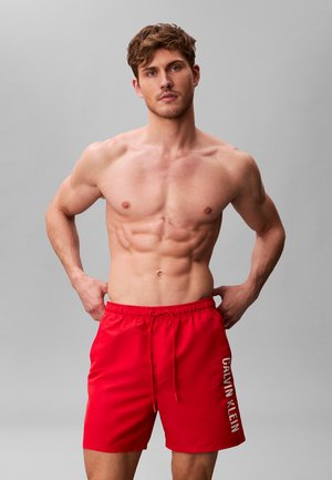 Muskuløs, bar overkrop mand iført røde Calvin Klein badeshorts står med hænderne på hoften mod en ensfarvet grå baggrund.