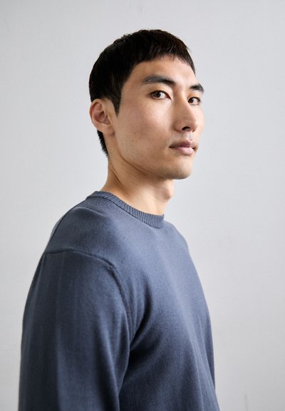 Pull en maille bleu avec un col rond et un tissu texturé, porté par une personne faisant légèrement face sur le côté contre un fond neutre.