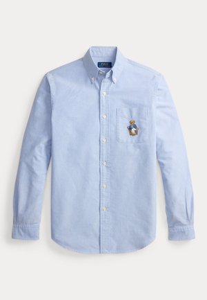 Chemise bleu clair à boutons, manches longues, avec une poche poitrine ornée d’un ours brodé portant un blazer bleu et un pantalon blanc.