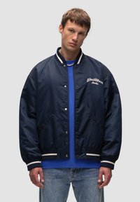 Granatowa kurtka bomber z gładką fakturą, białymi, prążkowanymi mankietami i wyszywanym logo. Noszona na niebieskiej koszuli i dżinsach.