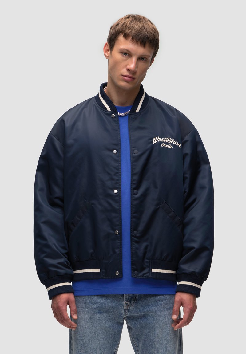Granatowa kurtka bomber z gładką fakturą, białymi, prążkowanymi mankietami i wyszywanym logo. Noszona na niebieskiej koszuli i dżinsach.