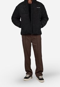 Cappotto imbottito in corduroy nero con zip frontale e tasche a toppa, abbinato a pantaloni marroni su misura e sneakers nere.