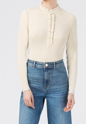 Bodysuit à manches longues couleur crème avec un col à volants et des détails de boutons, associé à un jean en denim bleu taille haute. Texture lisse et design ajusté.