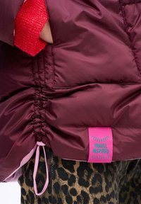 Burgundowa pikowana kurtka z troczkiem u dołu, z różową metką z napisem "INSPIRACJA PODRÓŻAMI" i spodniami w leopardzi wzór poniżej.