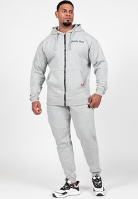 Grijze rits-hoodie met een capuchon met trekkoord, voorzakken en zwart logo. Gecombineerd met bijpassende grijze joggingbroeken. Witte en zwarte sportschoenen.