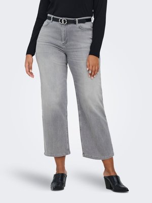 Femme portant un jean large gris clair raccourci, une ceinture noire avec une boucle argentée, un haut noir à manches longues, des mules noires à talons et des bagues argentées.