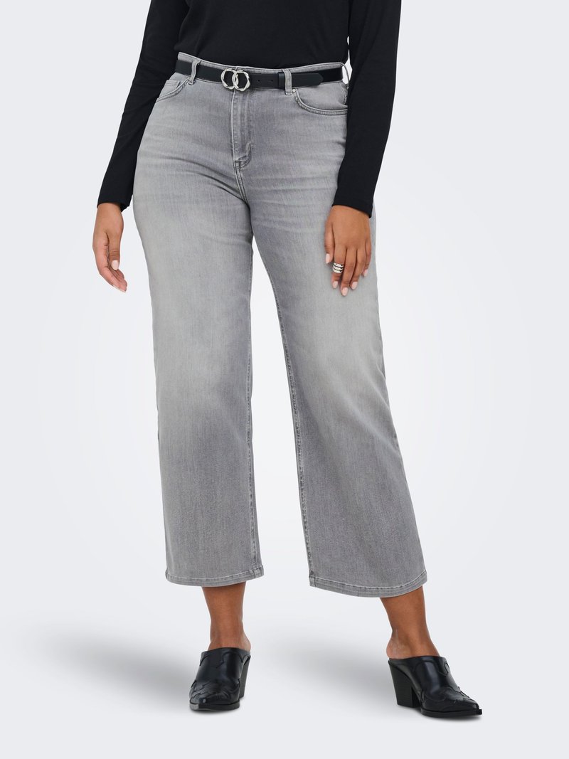 Femme portant un jean large gris clair raccourci, une ceinture noire avec une boucle argentée, un haut noir à manches longues, des mules noires à talons et des bagues argentées.