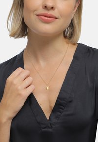 Collier pendentif doré avec un charme rectangulaire, présentant une surface lisse sur une chaîne fine, porté sur un haut noir à décolleté en V profond.