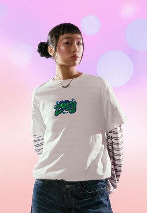 Fehér pamut póló zöld és lila graffiti stílusú "ZOEY" felirattal, fekete-fehér csíkos hosszú ujjú póló fölött viselve.