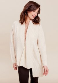 Beige open-front cardigan, textiele structuur, lange mouwen, zigzag patroon, zachte afwerking, valt tot aan de heupen, gedragen over een lichte top en zwarte broek.