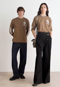 Brown long-sleeve shirts with white graphic accents, paired with dark denim and wide-leg black trousers. Casual footwear completes the look.