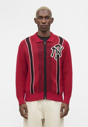 Man with short blonde hair wears red knit zip-up sweater with black and white vertical stripes and beige pants against plain white background.