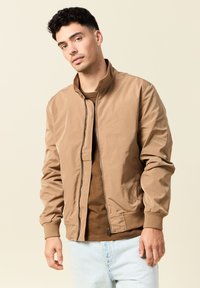 Chaqueta bomber marrón hecha de un tejido suave, con cierre de cremallera al frente, puños acanalados y cuello alzado, utilizada sobre una camiseta marrón y unos jeans claros.