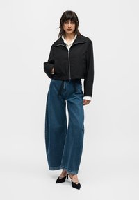 Γυναίκα με μαύρο μπουφάν με φερμουάρ, λευκό πουκάμισο, φαρδιά μπλε τζιν παντελόνια και μαύρα μυτερά τακούνια, στέκεται μπροστά σε λευκό απλό φόντο.