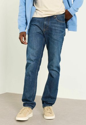 Mann trägt blaue Jeans, beige Turnschuhe, ein weißes T-Shirt und eine hellblaue Kapuzenjacke mit Reißverschluss, steht vor einem einfarbigen Hintergrund.