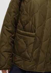 Casaco acolchoado verde oliva com um padrão texturizado em ondas, apresentando um bolso de remendo na parte da frente e um acabamento de tecido suave e liso.