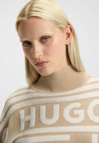 Beige en crèmekleurig gestreepte trui met een hoge ribbelkraag. Kenmerkt zich door het woord "HUGO" prominent in zachte, gestructureerde stof.