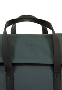 Bolsa impermeable verde con correas negras, con logotipo "RAINS" en relieve, textura suave y detalles en las costuras. Cierre con cremallera y botón a presión.