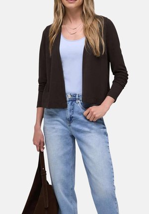 Mujer que lleva un cárdigan negro abierto, camiseta sin mangas azul claro, vaqueros de lavado claro, sosteniendo un bolso tote marrón oscuro a su lado.