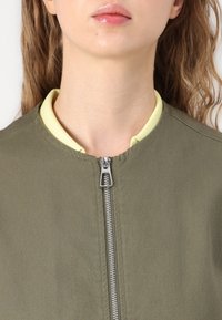 Olivgrüne Bomberjacke mit silbernem Reißverschluss, rundem Ausschnitt und gelbem Innenkragen. Glatte Textur und minimalistisches Design.