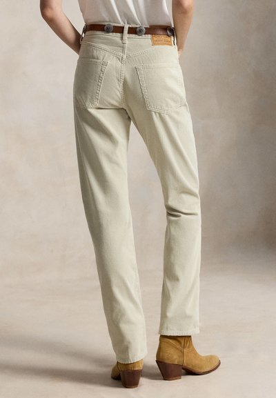 Personne portant un pantalon beige en velours côtelé droit avec poches arrière, ceinture marron et bottines couleur beige, debout sur un fond neutre.