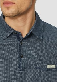Navyfarbendes, strukturiertes Poloshirt mit Button-Down-Kragen, Brusttasche und Etikettendetail. Es weist ein dezentes Muster und kontrastierende Knöpfe auf.