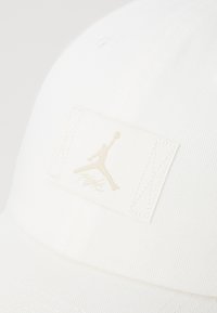Hvid kasket med et struktureret stof, et broderet logo-patch med en tan silhuet og teksten "Flight" i den nederste del af patchen.