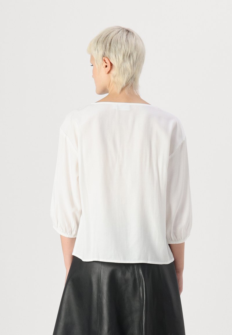 Blouse blanche semi-transparente à manches trois-quarts, col rond et ourlet légèrement court. Texture lisse, design minimal sans motifs.